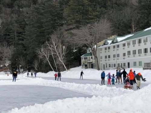 People ice skating on a frozen lake, with snowbanks and a lodge in the background surrounded by trees.
