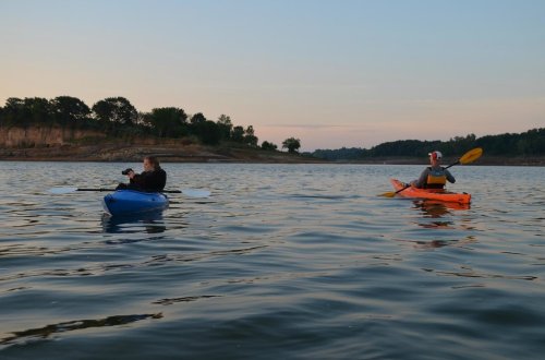 Two people kayaking on a calm lake during sunset, one in a blue kayak and the other in an orange kayak.