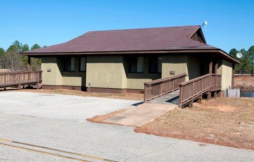 A single-story green building with a brown roof, featuring a ramp and surrounded by trees and a clear blue sky.