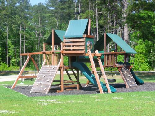 A wooden playground structure with slides, swings, and climbing features, set in a grassy area surrounded by trees.