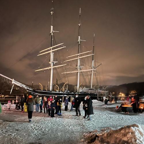 A crowd gathers near a tall ship in a snowy harbor at night, with lights illuminating the scene.