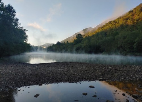 A serene river scene at dawn, with mist rising from the water and lush green hills in the background.