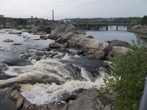 A river flows over rocky terrain, with a bridge and buildings visible in the background under a cloudy sky.