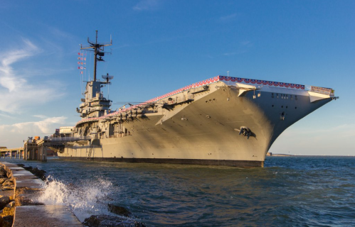 A large aircraft carrier docked at a pier, with flags and bunting along its deck, against a clear blue sky.