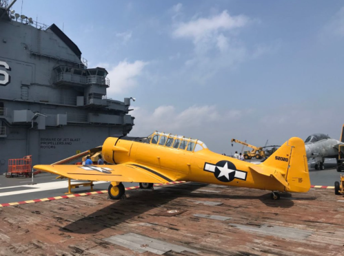 A yellow vintage aircraft on a flight deck, with a large ship in the background and a clear blue sky.