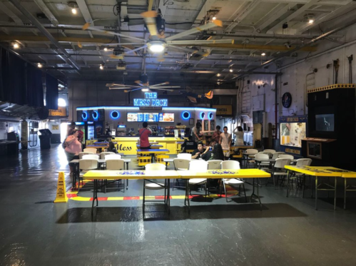Interior of a dining area with yellow tables and chairs, featuring a bar named "The Mess Deck" and people socializing.