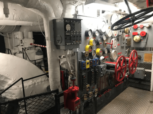 Control room of a ship featuring various gauges, valves, and machinery against a white backdrop.