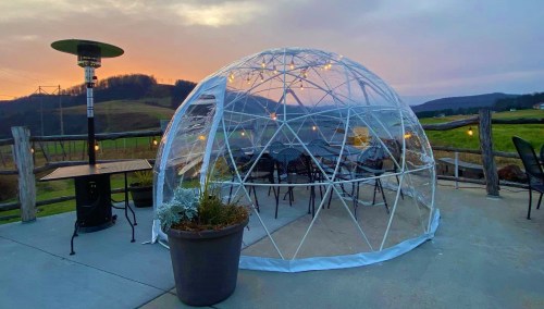 A clear geodesic dome with chairs inside, set against a sunset over rolling hills.
