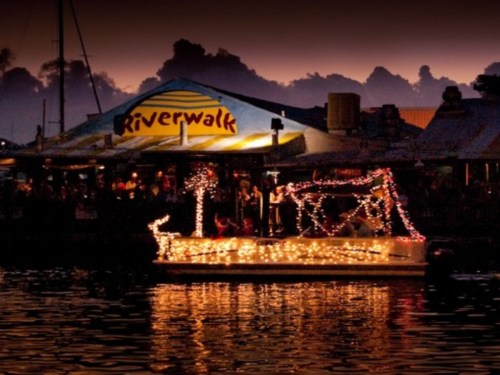A boat adorned with lights floats on water near a brightly lit Riverwalk restaurant at dusk.