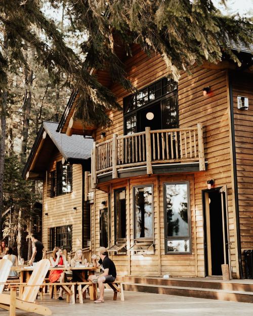 A wooden cabin surrounded by trees, with people sitting outside on a patio enjoying the outdoors.
