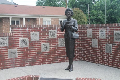 A bronze statue of a woman holding a book, surrounded by brick walls with plaques honoring notable individuals.
