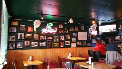 A cozy restaurant interior featuring a wall of framed photos, a neon sign, and tables with red chairs.
