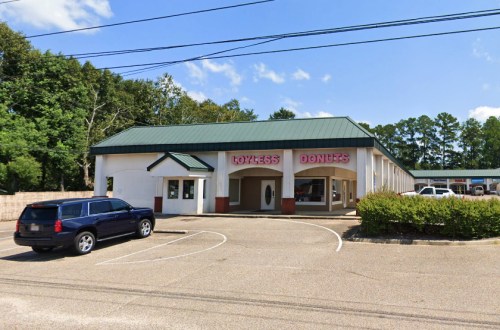 A donut shop with a green roof and pink signage, surrounded by trees and a parking lot.