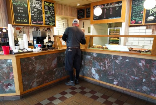 A person stands at a counter in a cozy café, ordering food with a menu displayed on the wall behind them.