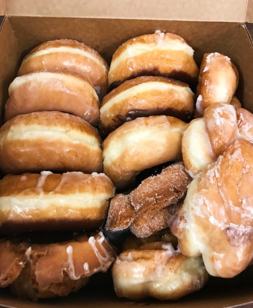 A box filled with assorted glazed donuts, including round and cruller shapes, some with sugar coating.