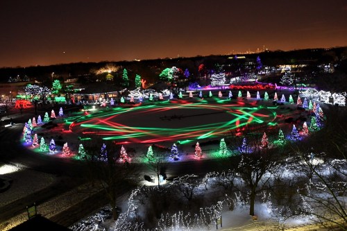 A festive park illuminated with colorful Christmas lights, featuring decorated trees and a circular light display at night.