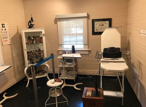 A vintage medical examination room with a chair, desk, microscope, and display cabinet filled with bottles.