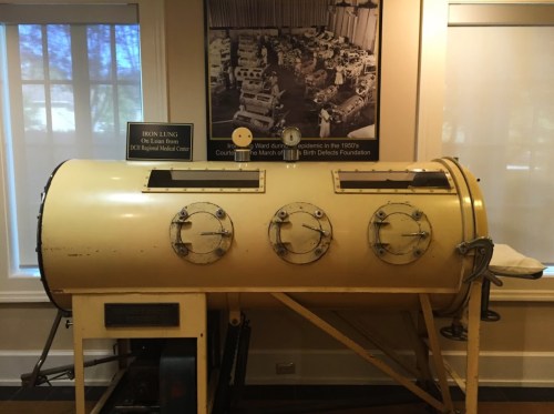 An iron lung machine displayed in a museum, with a historical photo of medical equipment in the background.