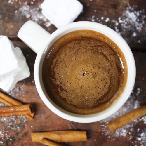 A white mug filled with coffee, surrounded by cinnamon sticks and sugar cubes on a wooden surface.