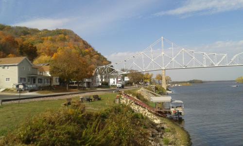 A scenic view of a river, bridge, and autumn-colored trees along the shore, with houses and a boat docked nearby.
