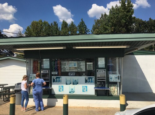 A small ice cream stand with two customers ordering, surrounded by trees and a blue sky with fluffy clouds.
