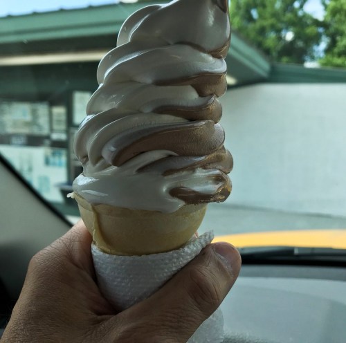 A hand holding a swirled chocolate and vanilla soft serve ice cream cone, with a blurred background of a building.