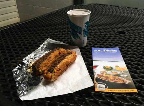 A hot dog in foil on a table with a drink and a brochure titled "100 Dishes to Eat in Alabama."