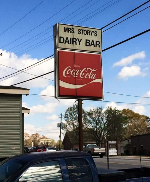 Sign for Mrs. Story's Dairy Bar featuring a Coca-Cola advertisement against a blue sky with clouds.