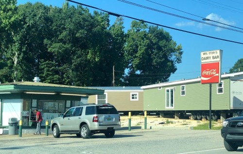 A roadside dairy bar with a sign, surrounded by trees and a parked SUV, with a mobile home in the background.
