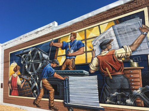 A colorful mural depicting workers in a factory setting, showcasing teamwork and industrial tools against a blue sky.