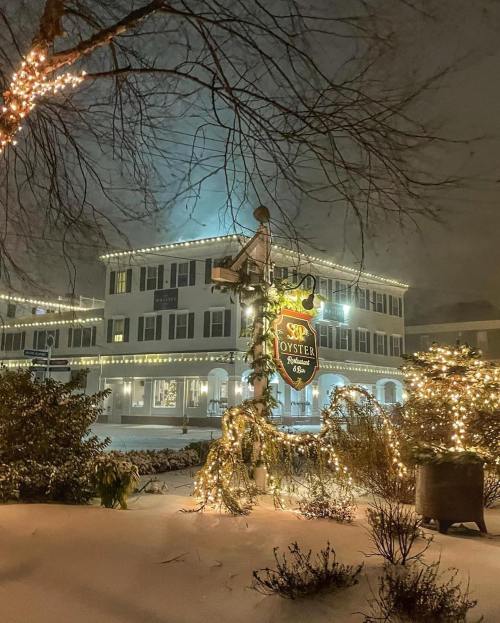 A snowy scene featuring a warmly lit restaurant sign surrounded by festive holiday decorations and trees.