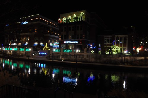 Colorful holiday lights adorn buildings along a canal at night, reflecting in the water.