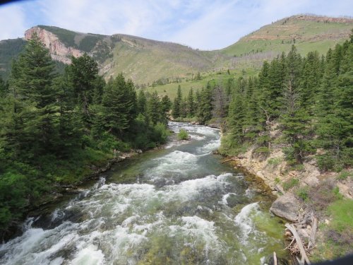 A flowing river surrounded by lush green trees and mountains under a blue sky with scattered clouds.