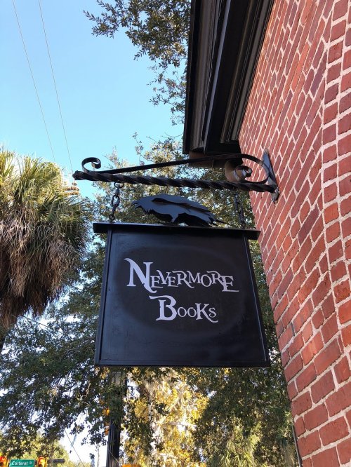 Sign for "Nevermore Books" featuring a raven silhouette, hanging from a decorative bracket against a brick wall.