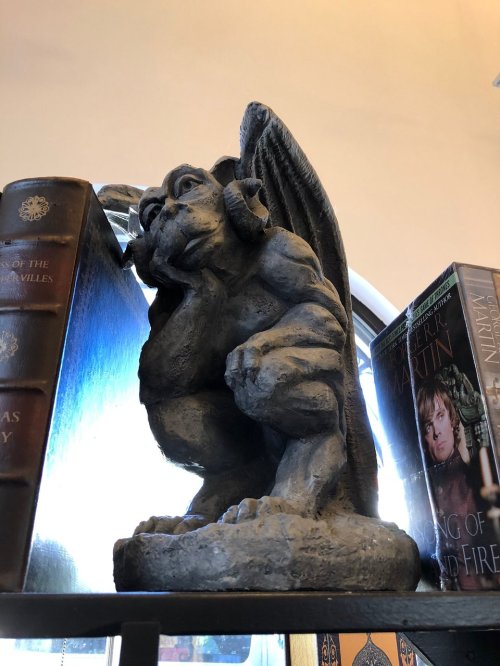 A stone gargoyle statue with wings, perched on a shelf beside books.