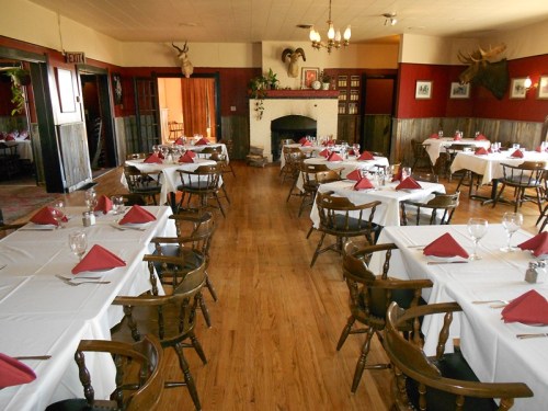 A cozy restaurant interior with wooden tables set for dining, featuring white tablecloths and red napkins, and a fireplace.