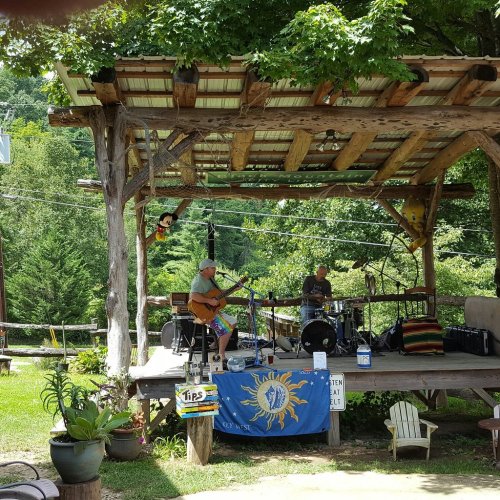 Two musicians perform on a rustic outdoor stage surrounded by greenery, with a colorful banner and plants nearby.