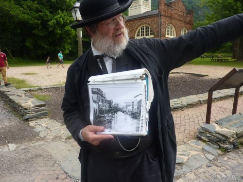 A man in period clothing points while holding a binder with a historical photo of a flooded street.