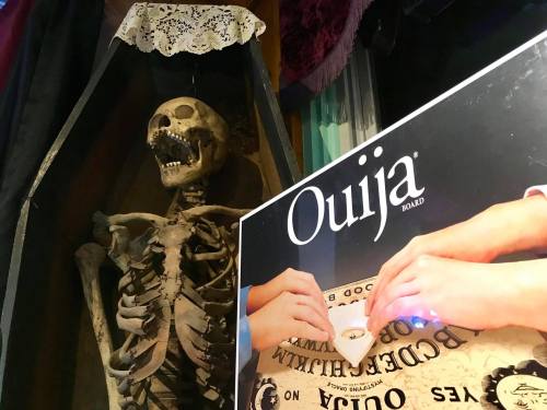 A skeleton in a coffin next to a Ouija board with hands poised over the planchette.