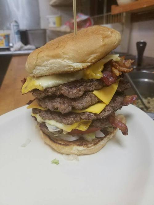 A towering burger stacked with multiple beef patties, cheese, bacon, lettuce, and tomato on a toasted bun.