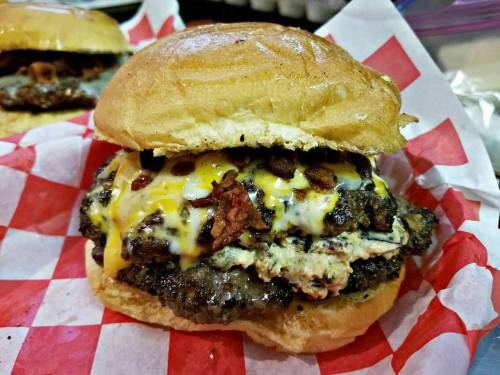A close-up of a juicy burger with melted cheese, bacon, and a toasted bun, served on red and white checkered paper.