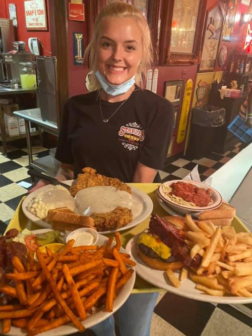 A smiling waitress holds a large tray of fried chicken, sides, and a burger in a retro diner setting.
