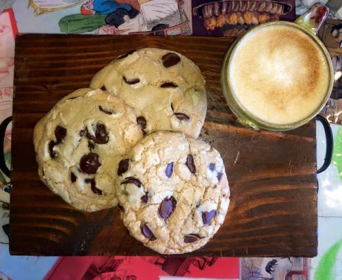 Three chocolate chip cookies on a wooden board next to a cup of frothy coffee. Colorful background.