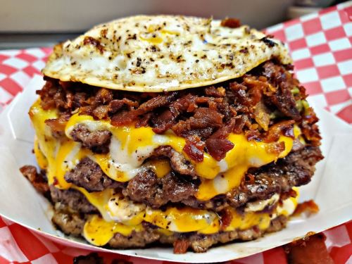 A towering burger with multiple beef patties, melted cheese, crispy bacon, and a fried egg on top, served in a paper tray.