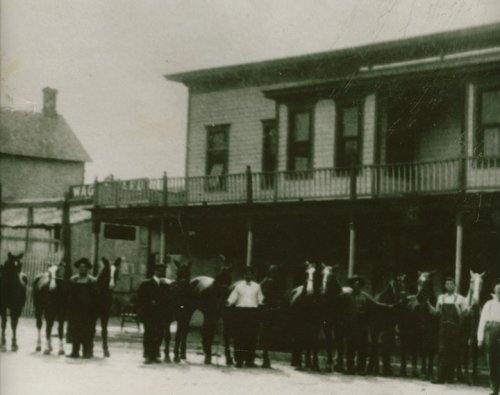 Black and white photo of a group of people with horses in front of a building, likely a general store or hotel.