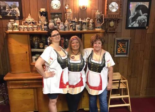 Three women in traditional dresses pose together in a cozy room filled with decorative items and wooden furniture.