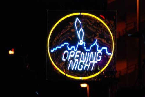 Neon sign reading "Opening Night" with a city skyline silhouette in the background.
