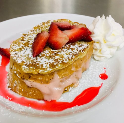 A slice of French toast layered with strawberry ice cream, topped with fresh strawberries, whipped cream, and a berry sauce.