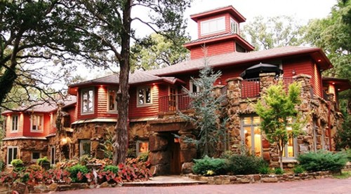 A large, red wooden house with stone accents, surrounded by trees and colorful flowers in a serene setting.