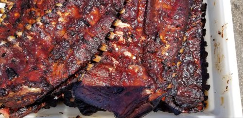 Close-up of grilled, smoky barbecue ribs with a rich, dark glaze, arranged in a white tray.